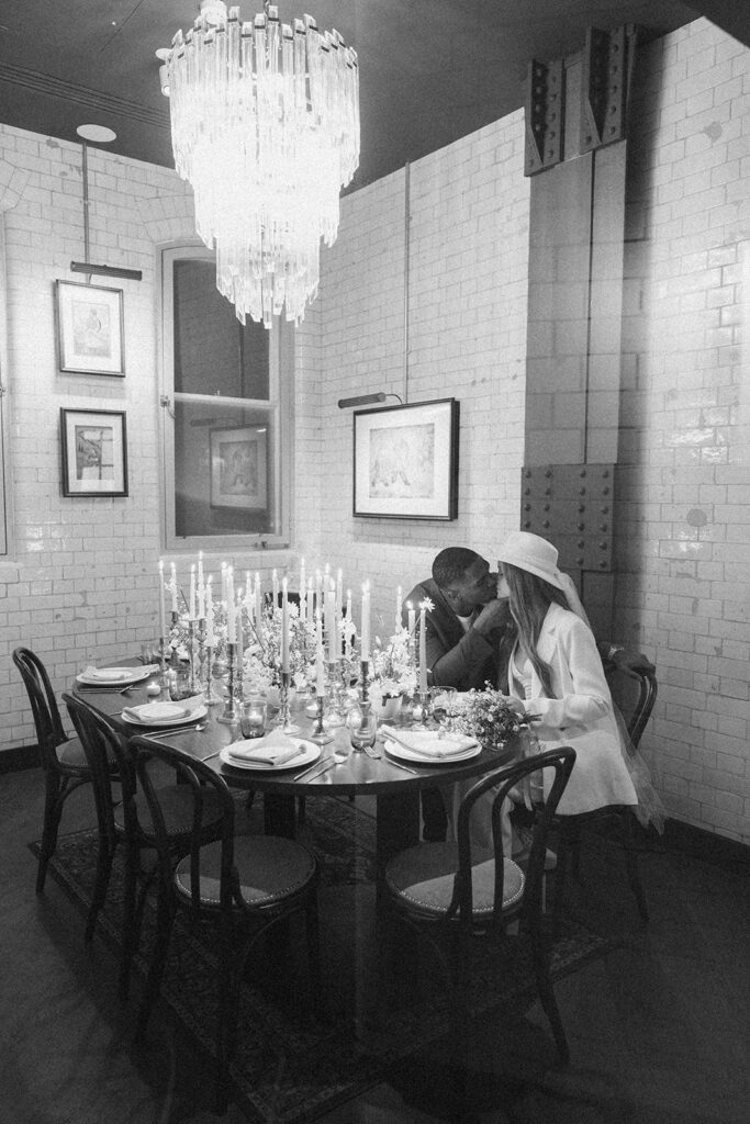 Black and white photo of groom kissing the bride at wedding dinner table